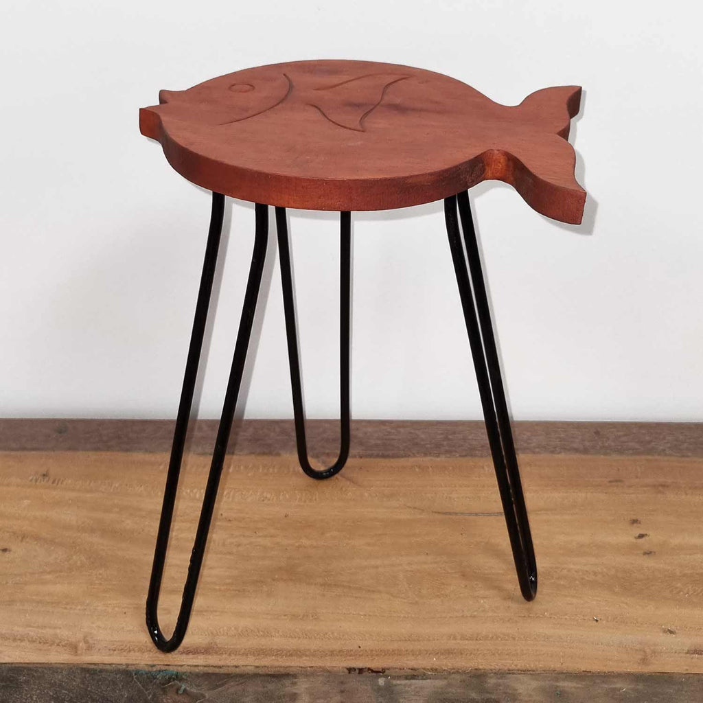 Wooden fish-shaped stool with black metal legs on a wooden floor.