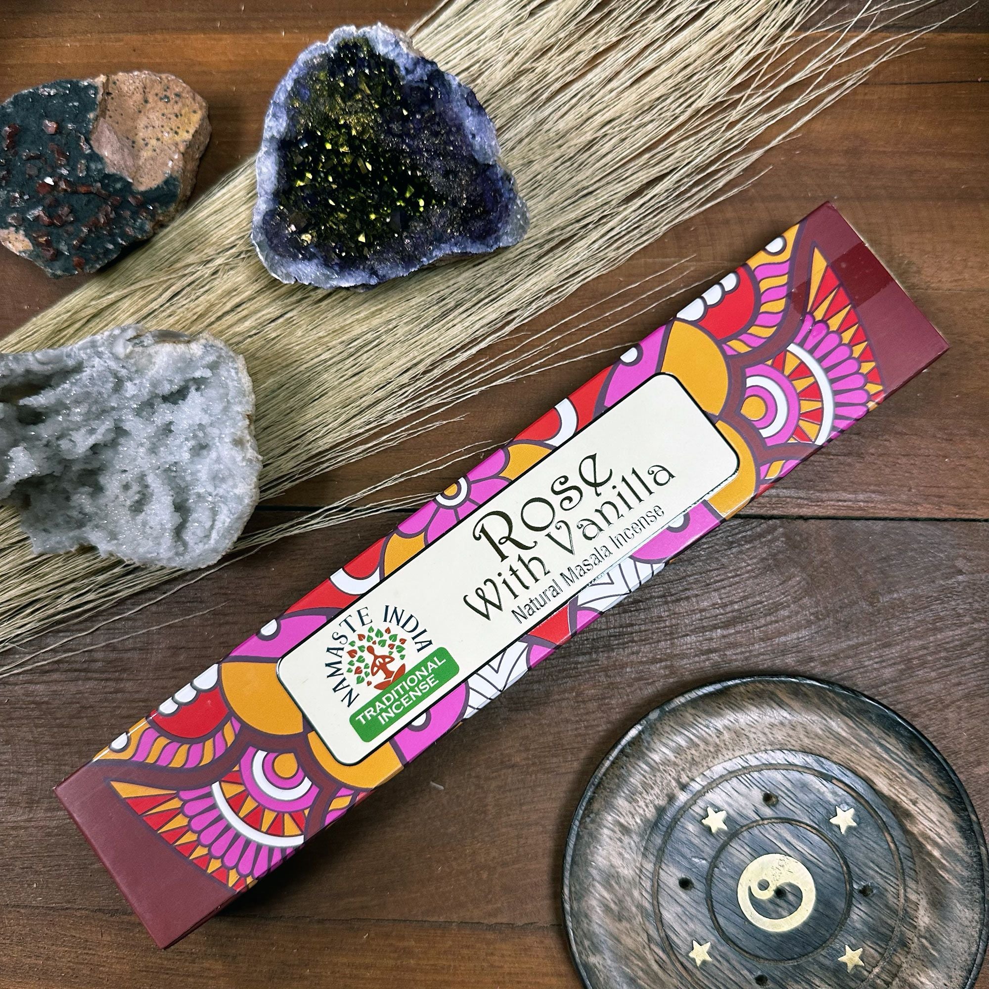 Colorful packaging of 'Rose with Vanilla' incense on a wooden surface with crystals and a decorative plate.