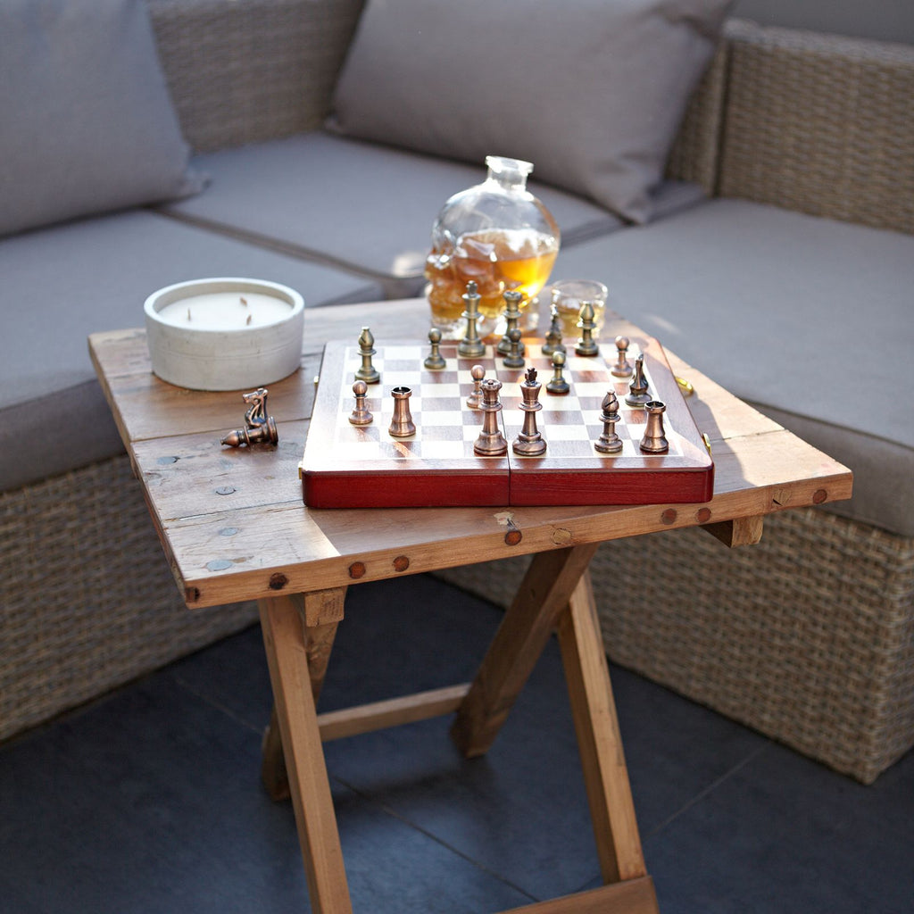 Wooden table with chessboard, glasses, and decanter on a patio setting.