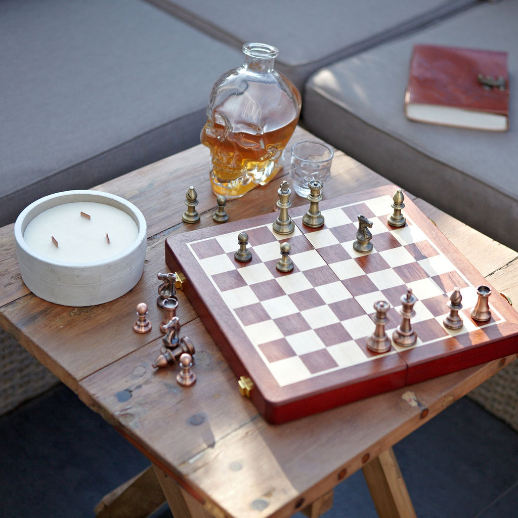 Chessboard with pieces, skull-shaped decanter, and candle on a wooden table.