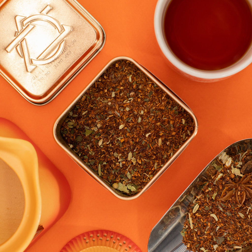 Spice tin with tea leaves, a spoon, and a cup of tea on an orange background