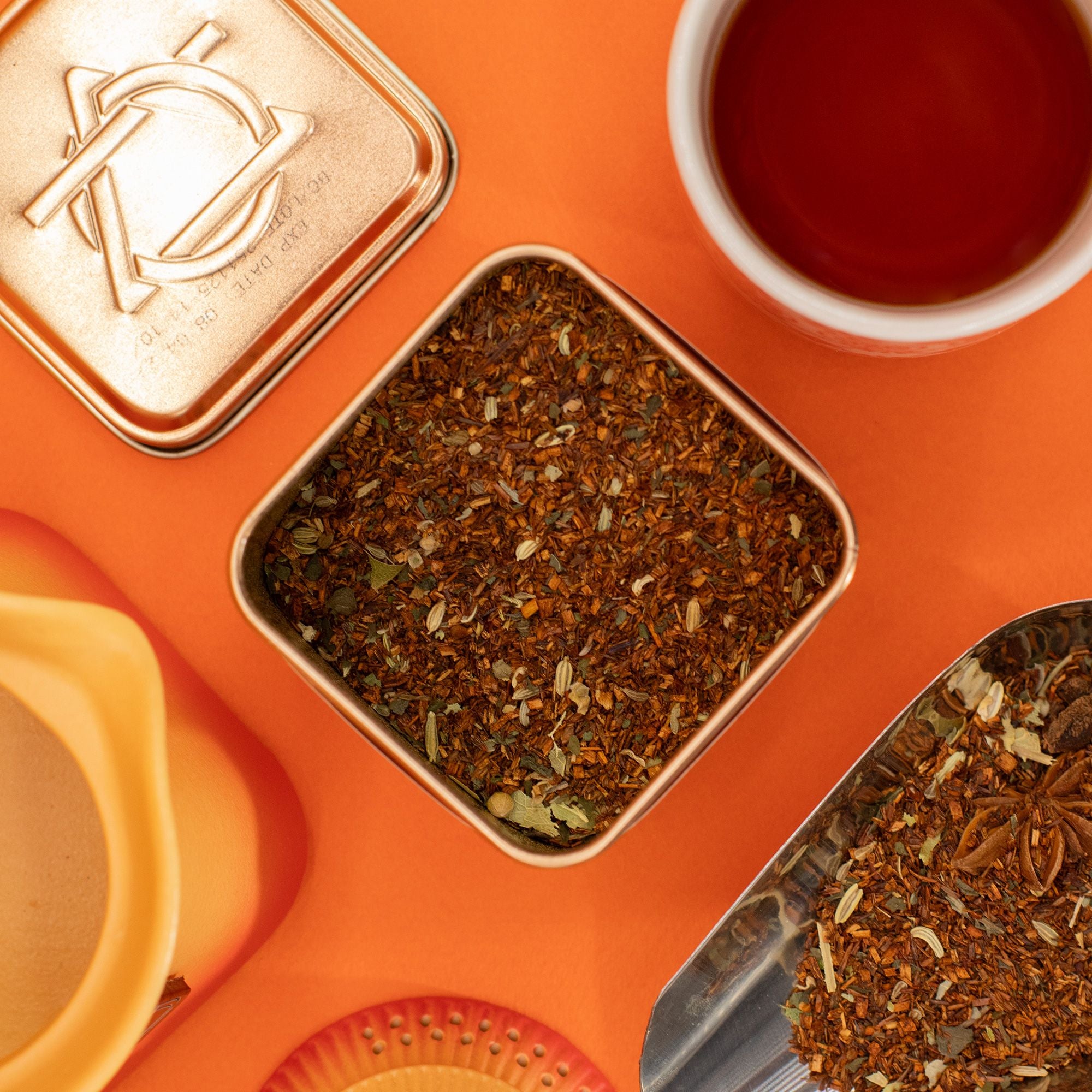 Spice tin with tea leaves, a spoon, and a cup of tea on an orange background