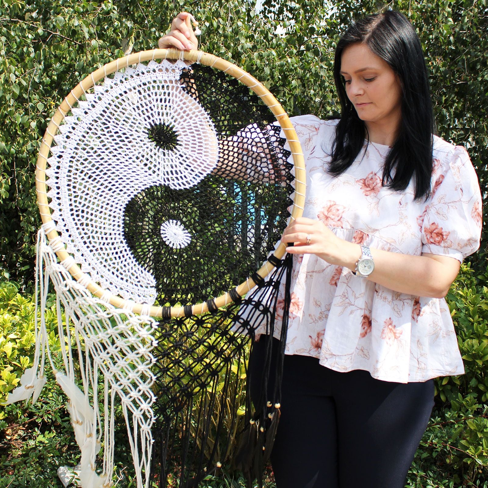 Woman holding a large macrame wall hanging with a yin yang symbol against a green outdoor background.