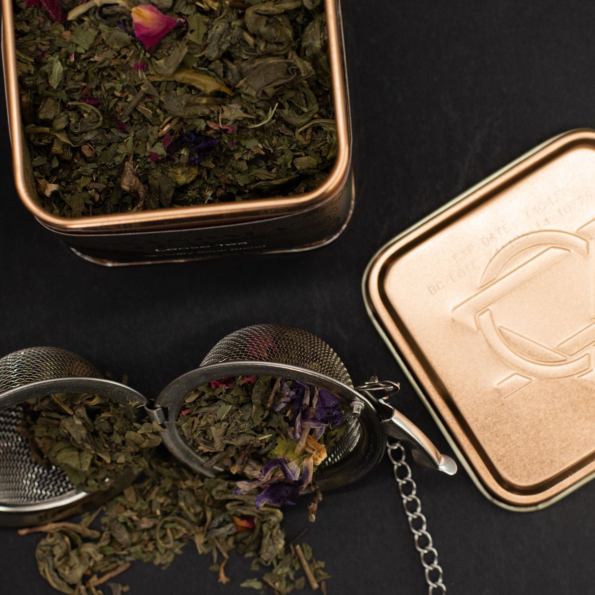 Tea leaves in a metal tin with a mesh strainer on a dark surface