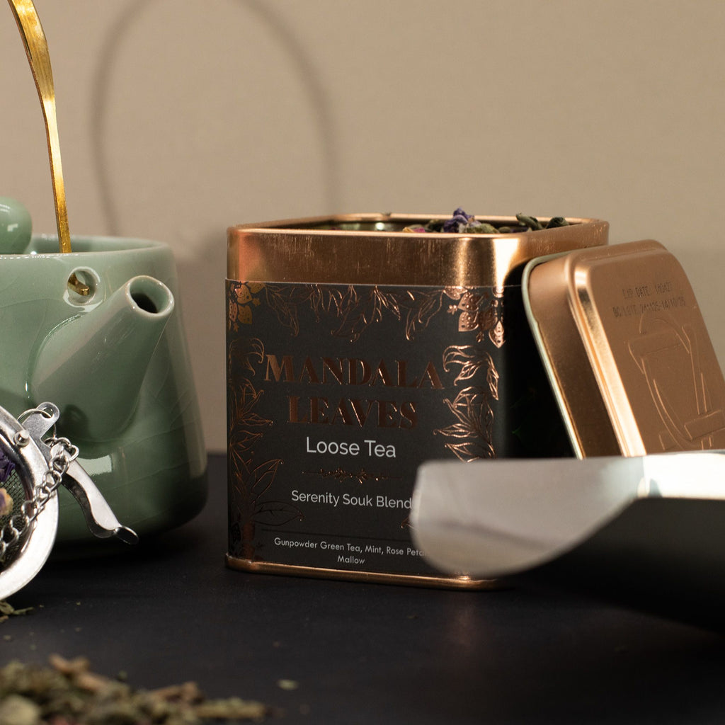 Tea canister with 'Mandala Leaves' branding next to a green teapot and silver tea strainer.
