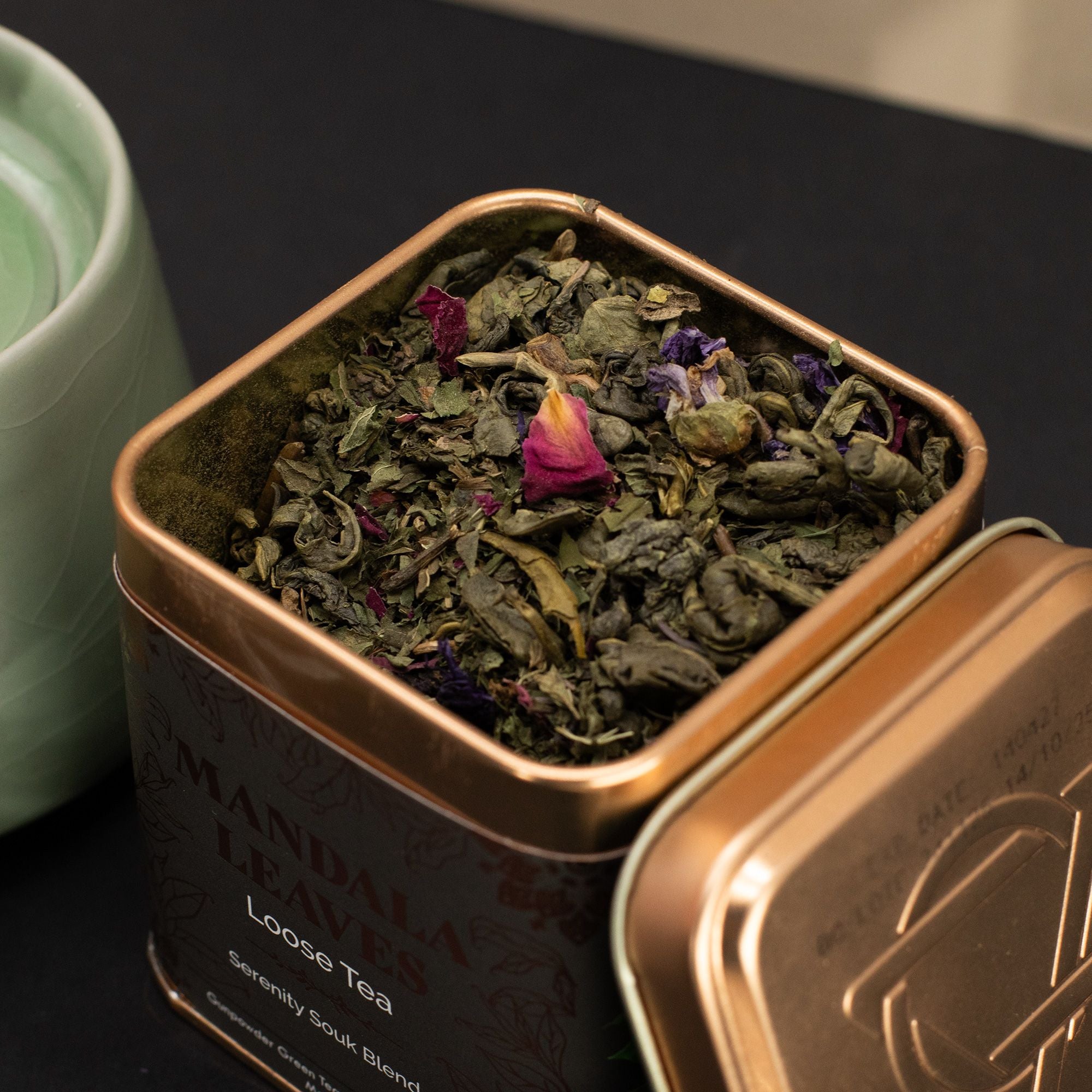 Open tin of loose tea with a green tea leaf and flower, next to a green teacup.