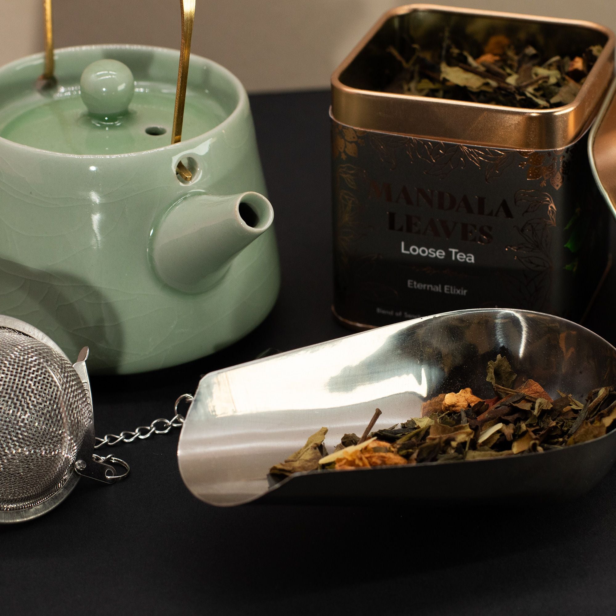 Tea-making set with green teapot, metal tea strainer, and container of loose tea leaves on a dark surface.