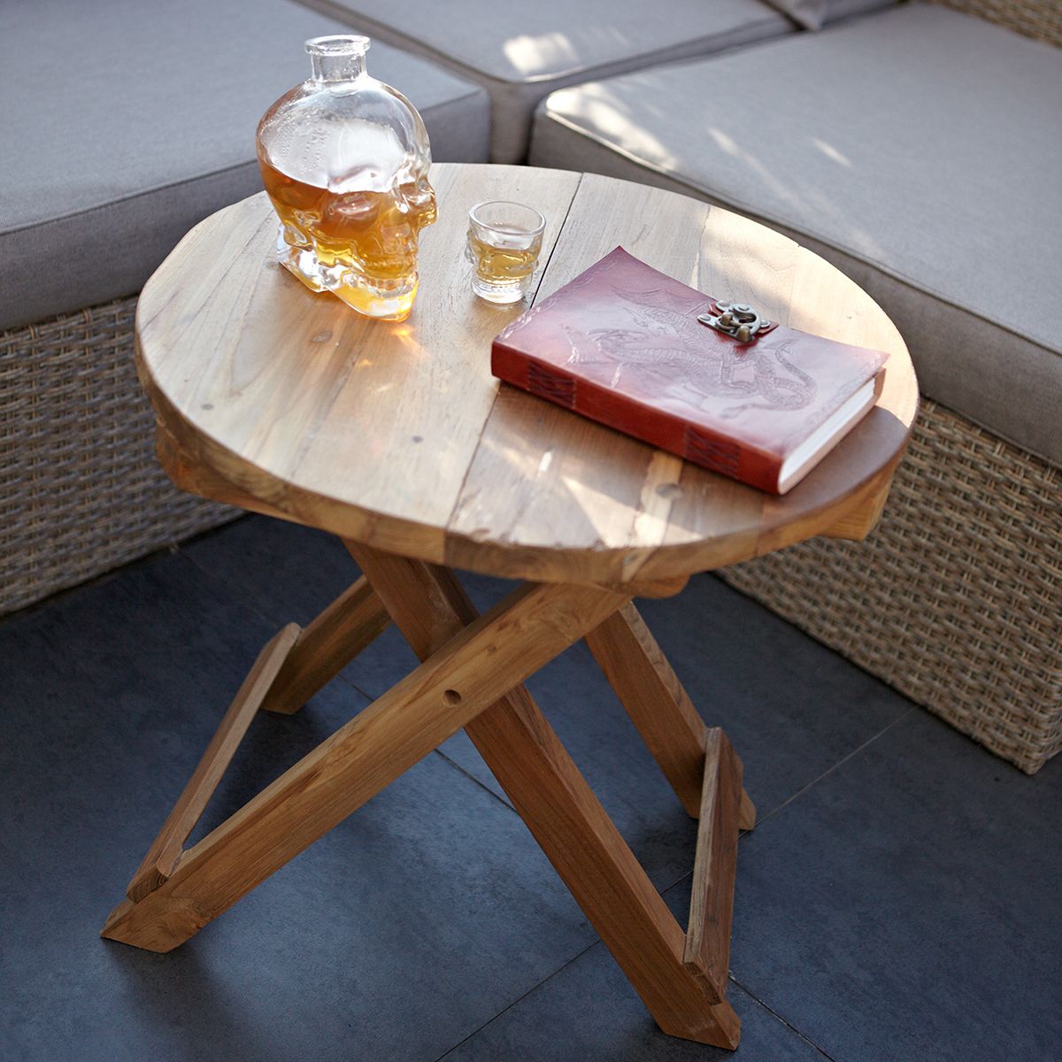 Wooden side table with a skull-shaped decanter, glass, and book on a patio.