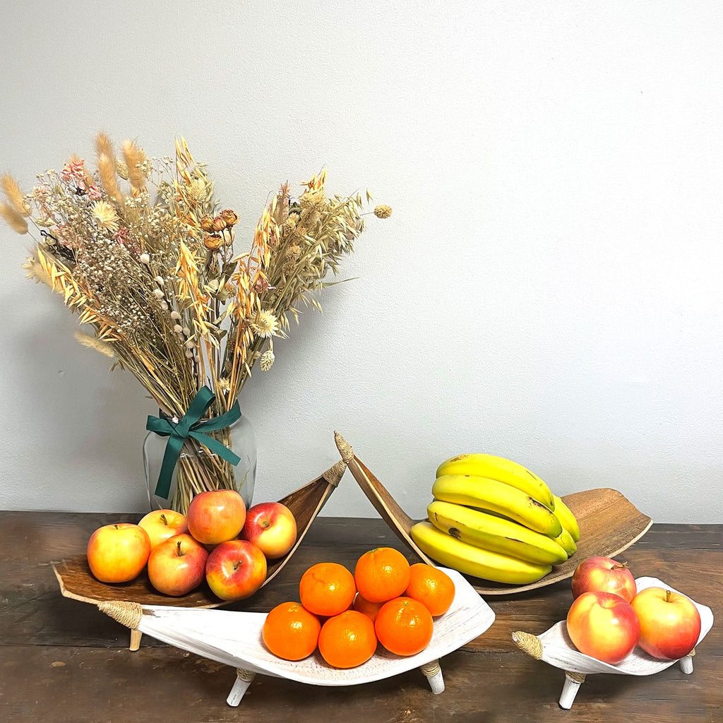 Fruit display with apples, oranges, bananas, and a vase of dried flowers on a wooden table.