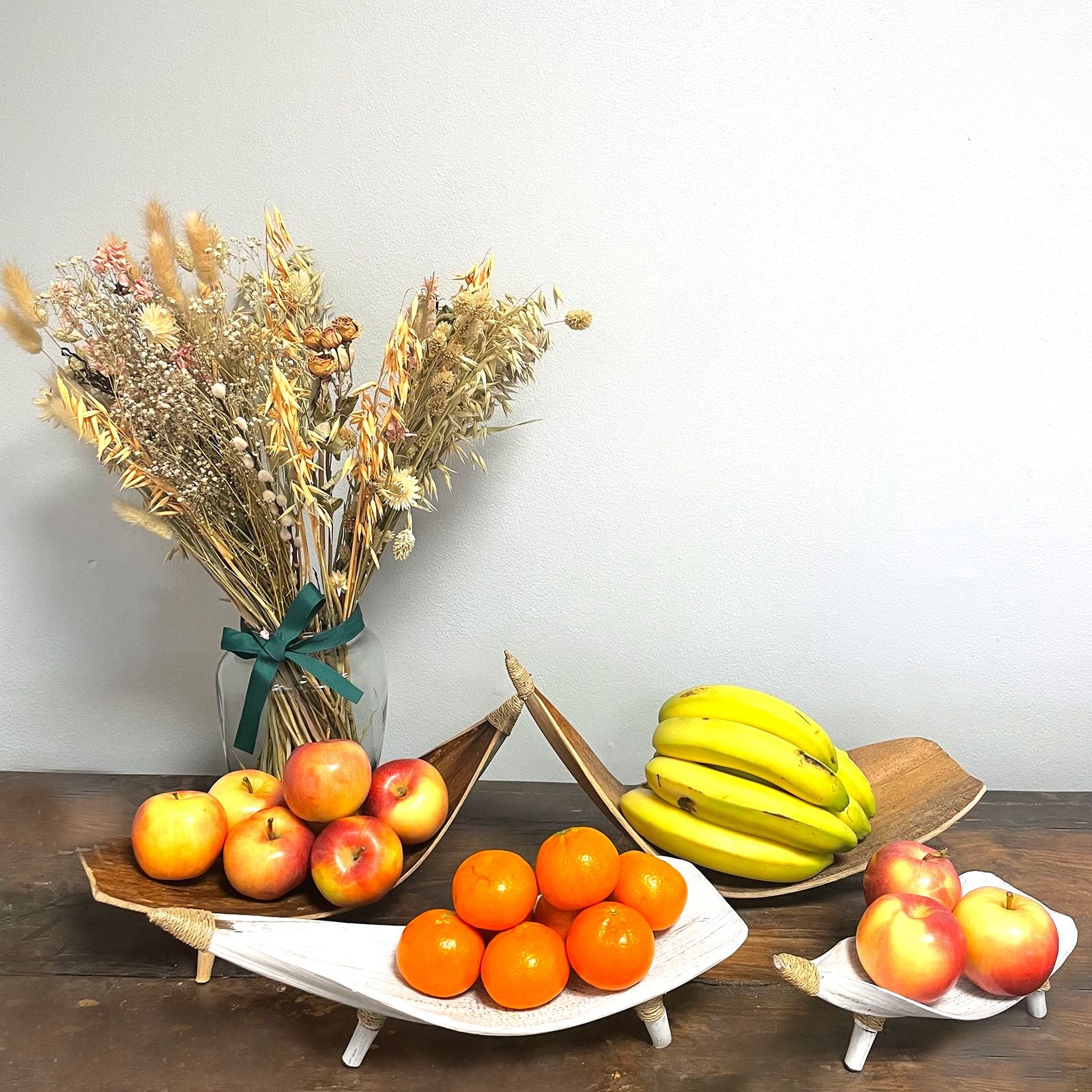 Fruit display with apples, oranges, bananas, and a vase of dried flowers on a wooden table.