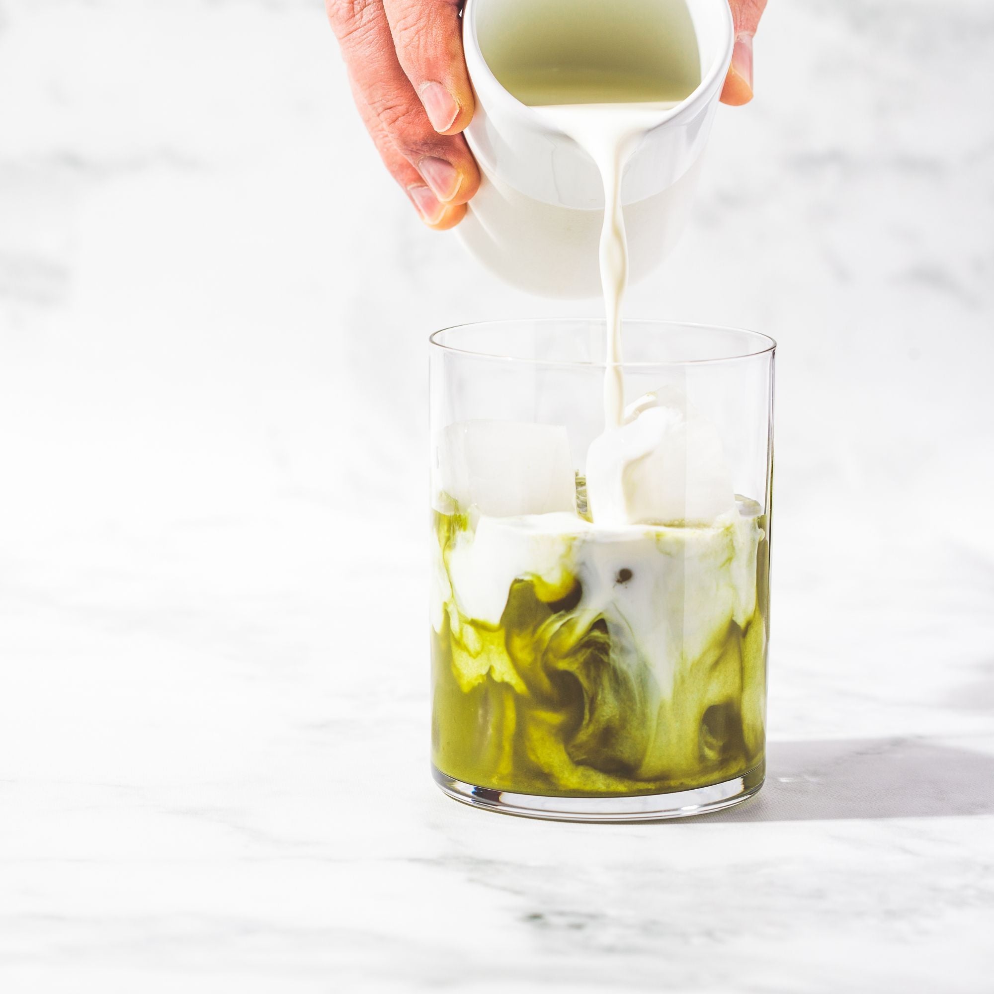 Hand pouring a white liquid into a glass with green liquid, on a white background