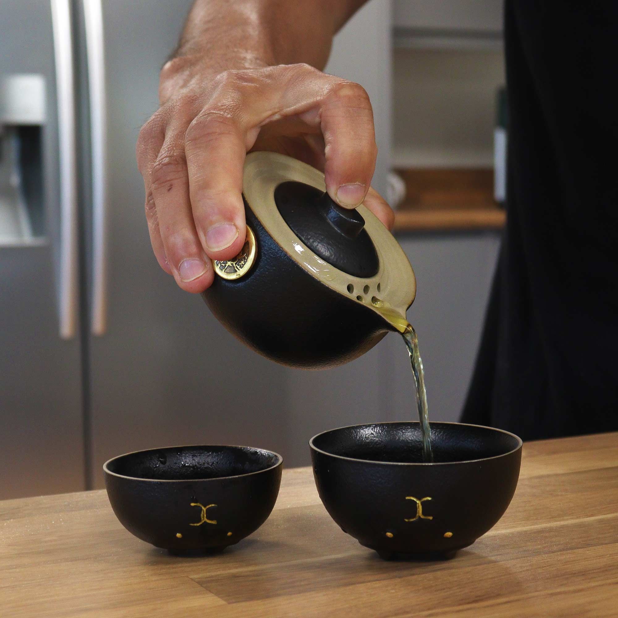 Person pouring tea from a black teapot into two black cups on a wooden surface.