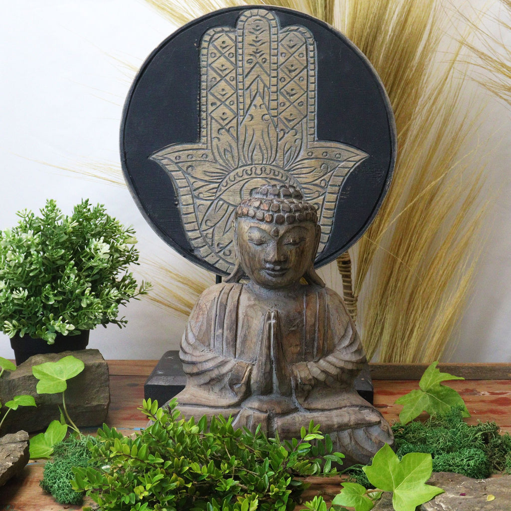 Wooden Buddha statue with Hamsa hand symbol in a decorative setting with plants and stones.