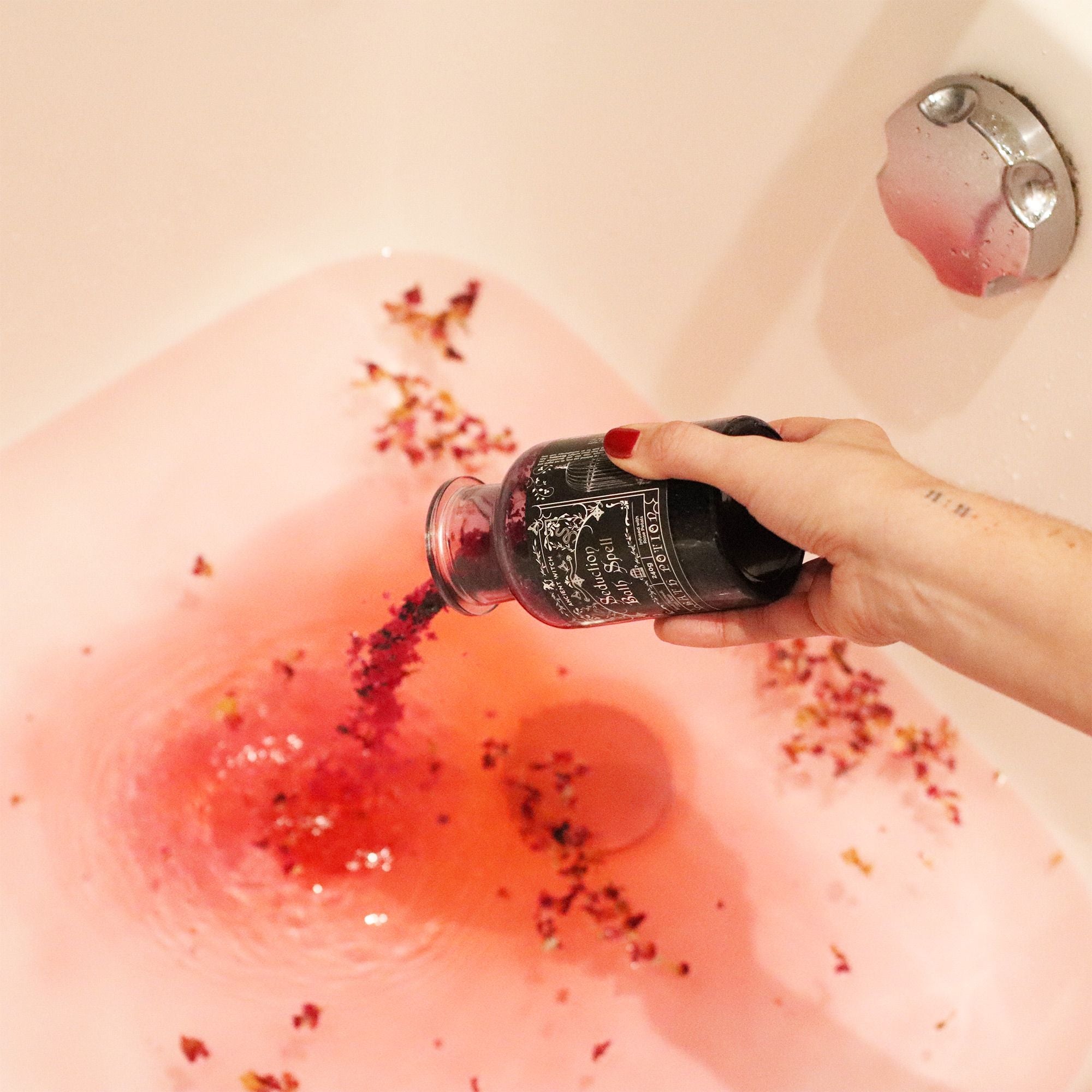 Hand pouring red liquid from a bottle into a bathtub filled with water and flowers.