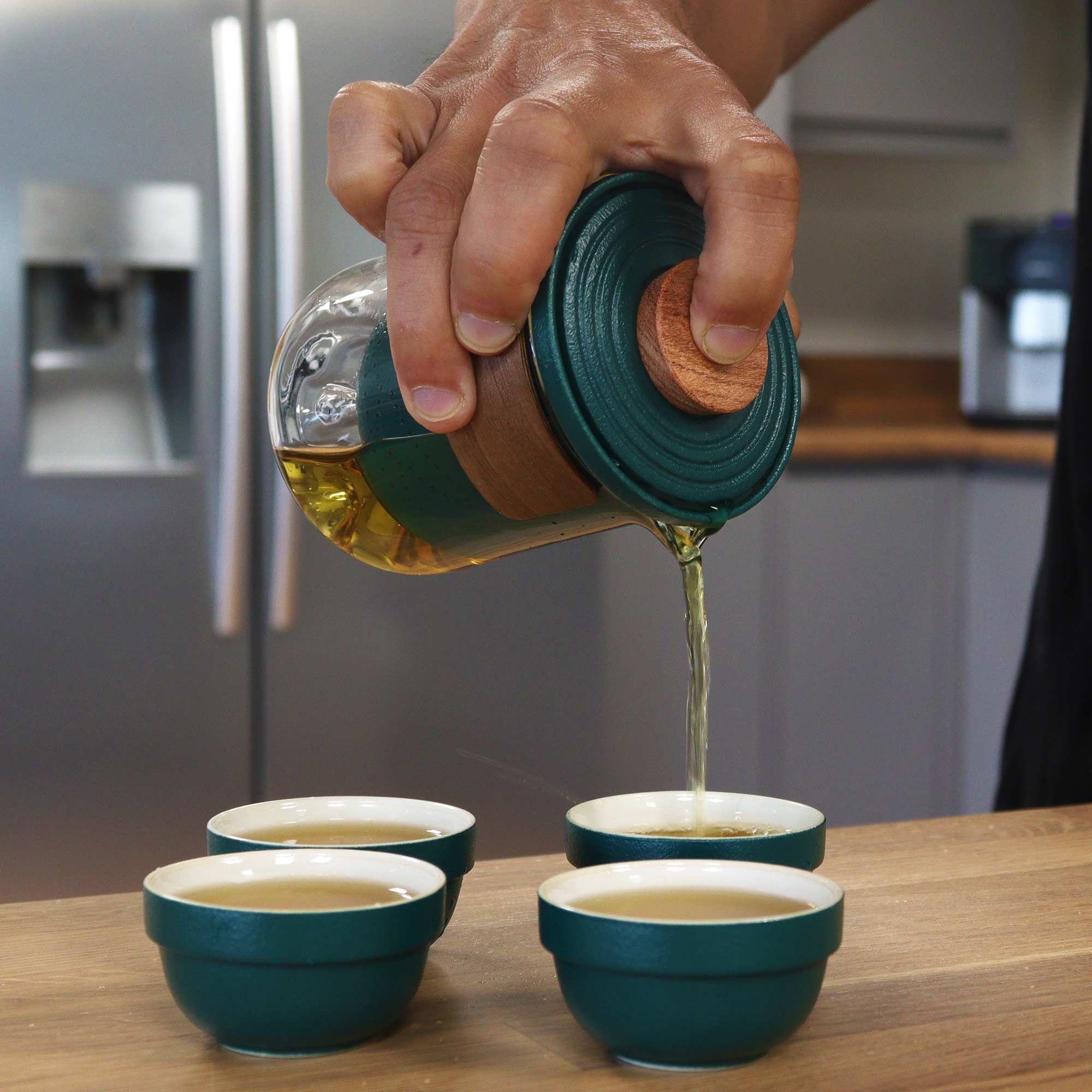 Hand pouring liquid from a green container into four small green bowls on a wooden surface.