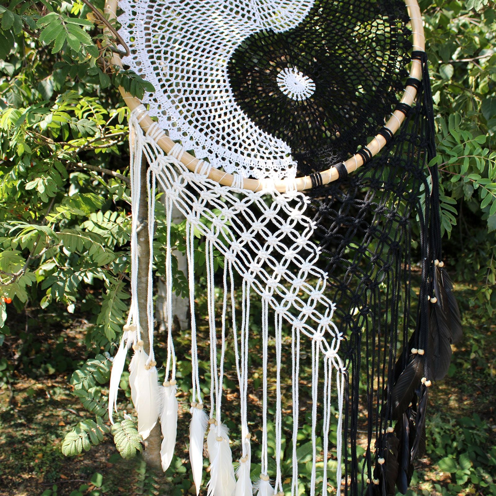 Macrame wall hanging with white and black yarn and feathers, set against a natural outdoor background.