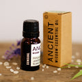 Bottle and box of Ancient Wisdom essential oil on a wooden surface with flowers in the background